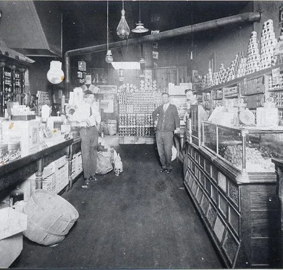 Early 1900s general store interior with stocked aisle, counters, glass display case, and shelves.