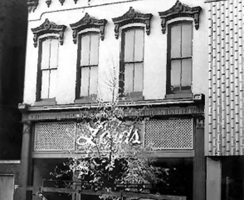 Historic black-and-white storefront with tree, script sign, and three upper windows on a street corner.