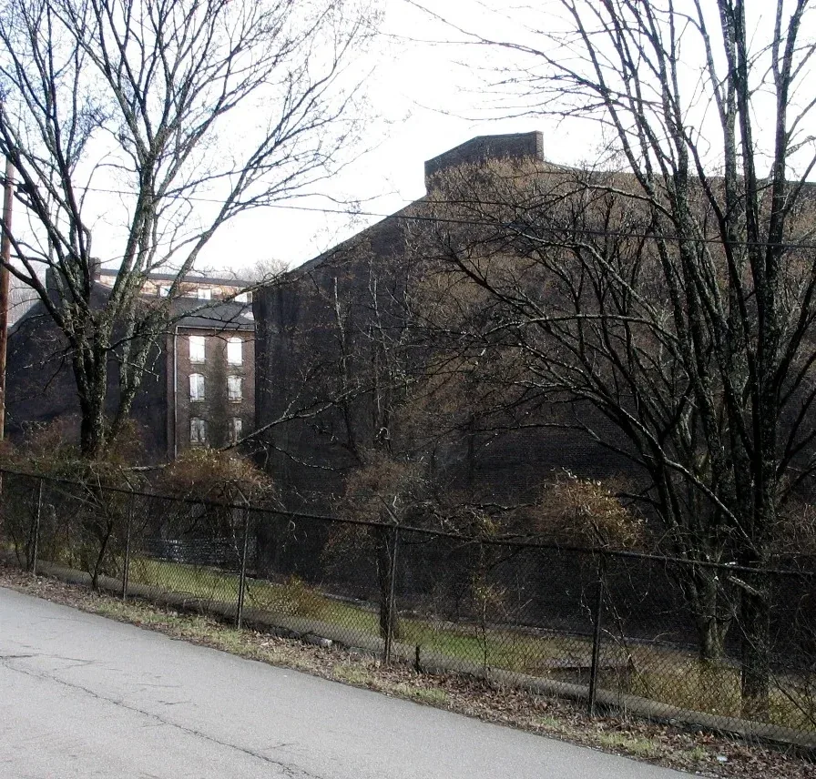 Abandoned brick industrial wall beside overgrown chain-link fence along damp winter road, quiet gray landscape