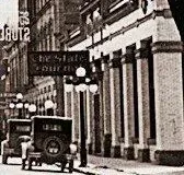 Vintage black-and-white downtown street view with parked early car, hanging sign, classic buildings.