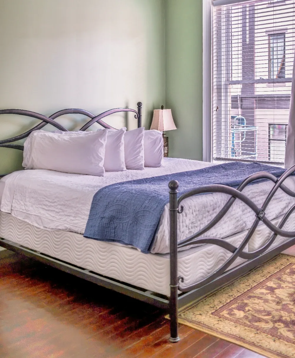 Sunlit green bedroom with white linens, blue throw, and metal lattice bed frame near window