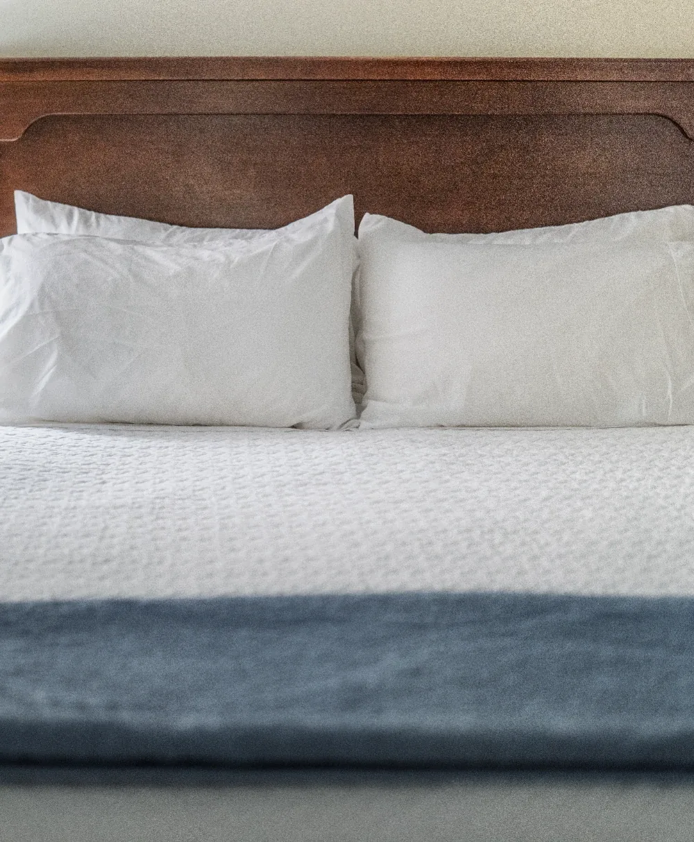 Minimal white bed with pillows and warm wooden headboard, soft textured linens, blue throw blanket