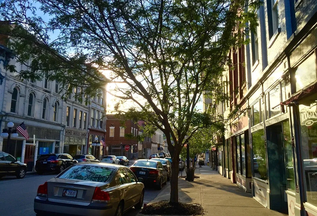 Historic brick storefronts along a canopy-shaded street with parked cars and warm afternoon light