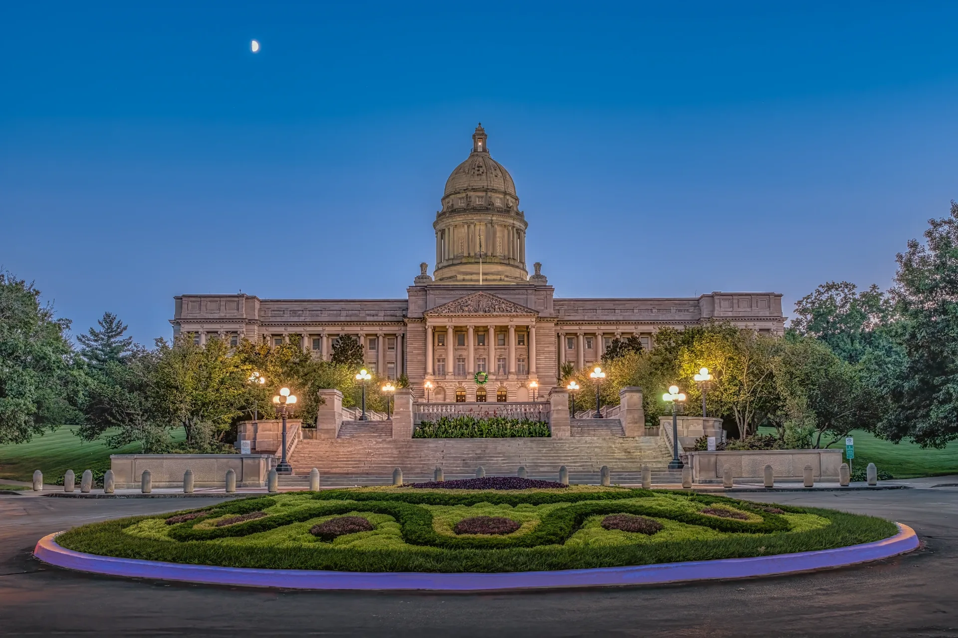 Neoclassical government dome at twilight with lit garden steps and manicured circular hedges