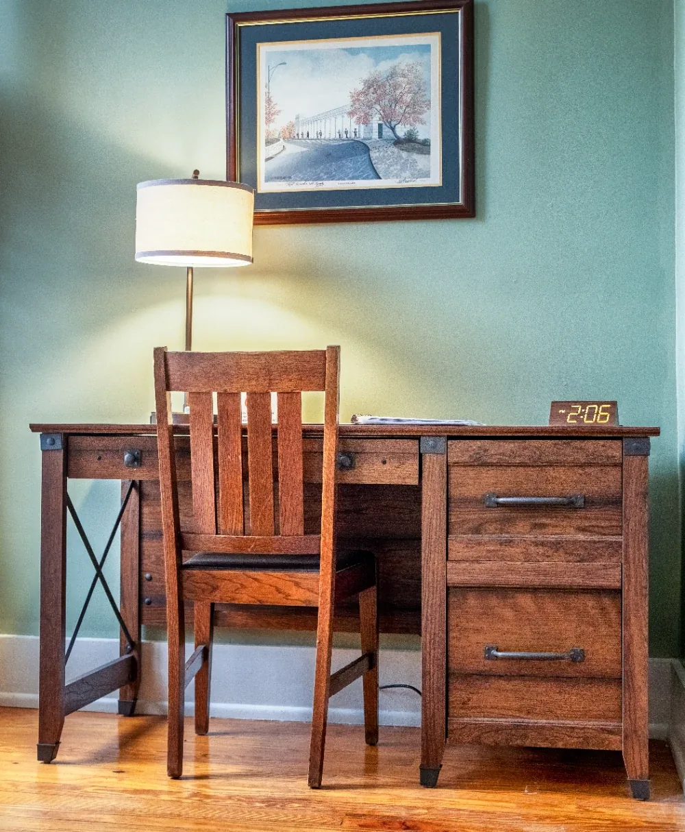 Warm home office mission desk with lamp, clock, framed landscape, and wooden chair.