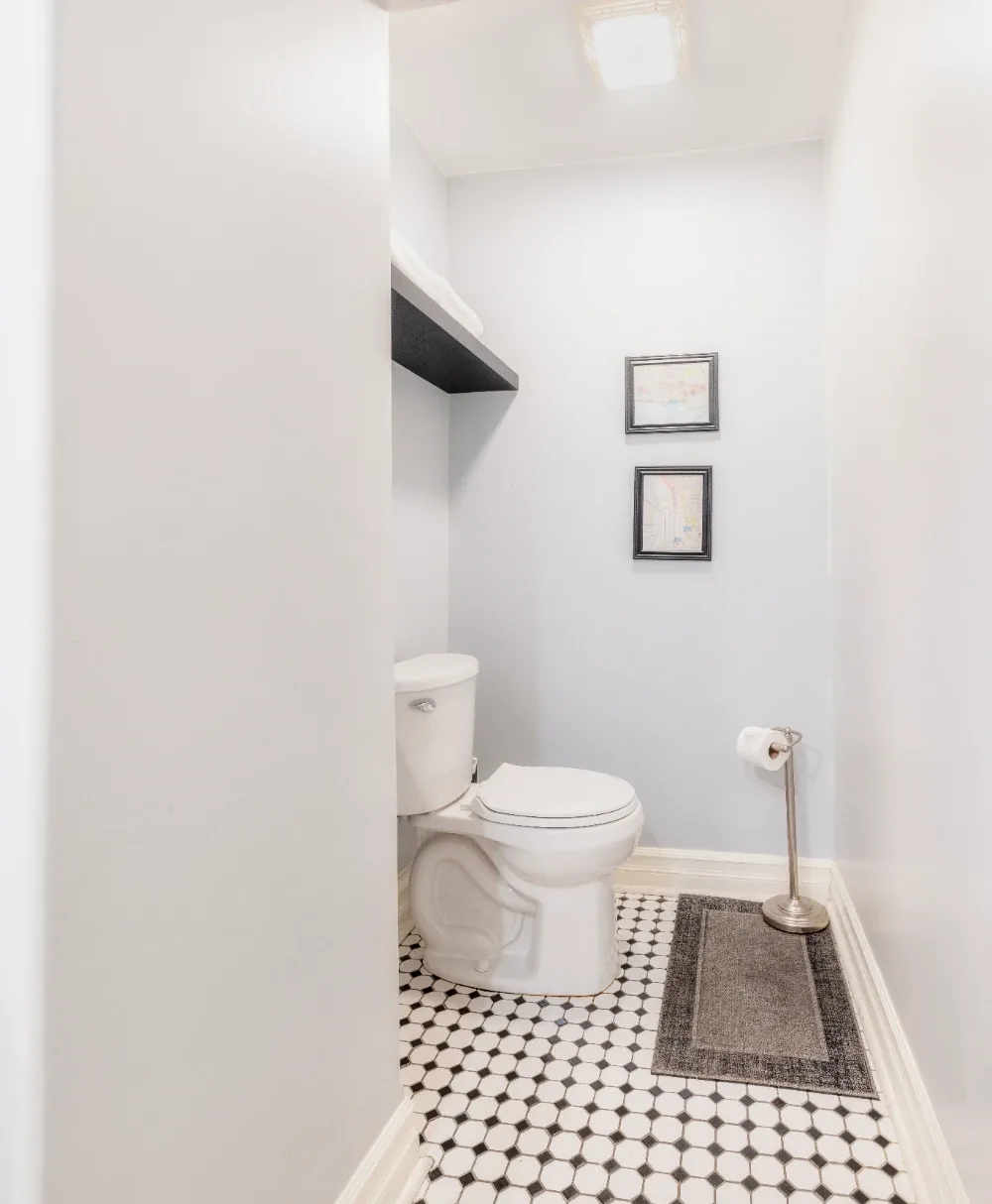 Minimal white small bathroom with floating shelf, framed art, and black-and-white tile toilet