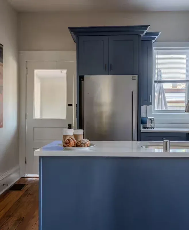 Modern blue kitchen island with pastries, stainless refrigerator, bright window, and hardwood floors