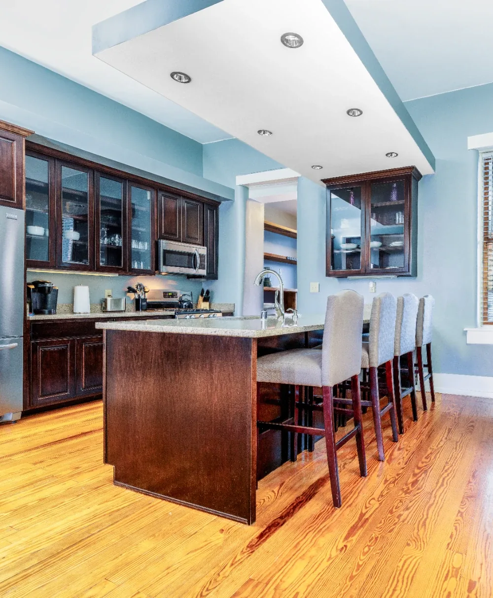 Modern espresso kitchen island with stone counter, seating stools, glass cabinets, and recessed lighting.