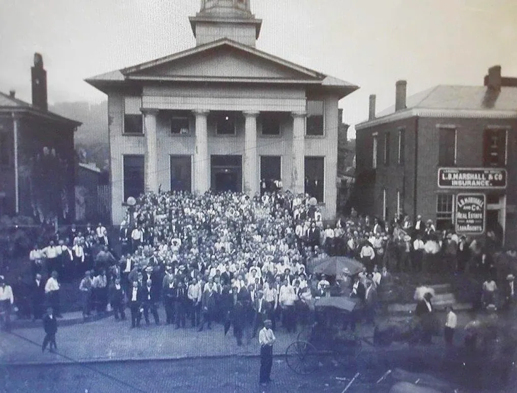 Historic town courthouse crowded grand steps, early cars, and period clothing in monochrome photo