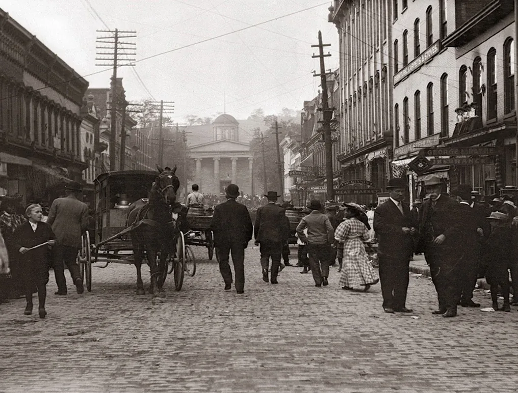 Historic Main Street with cobblestones, horse-drawn carriages, and period pedestrians near a classical building