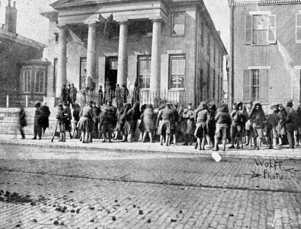 Civic crowd gathers outside historic courthouse with columns, steps, and attentive men in winter coats