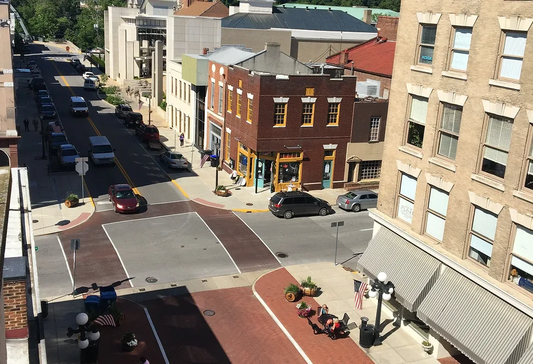 Sunny brick crosswalk downtown intersection with storefronts, cars, and pedestrians in bright daylight