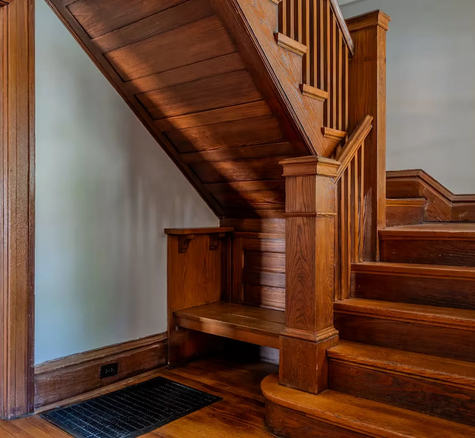 Honey-brown wooden staircase with built-in bench, paneled underside, handrail, and black floor vent cover