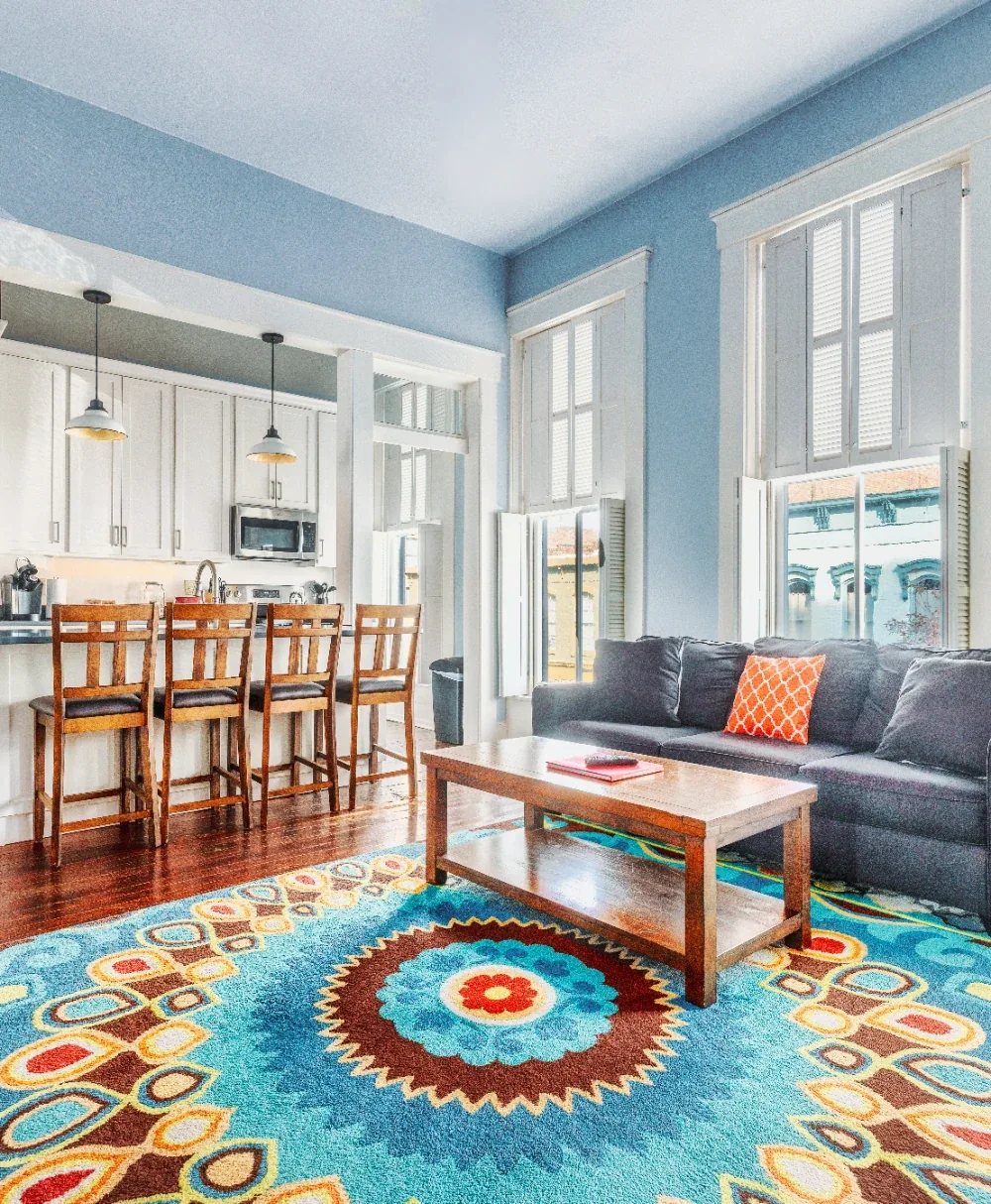 Sunlit coastal living room with mandala rug, blue walls, modern sofa, and white kitchen bar stools